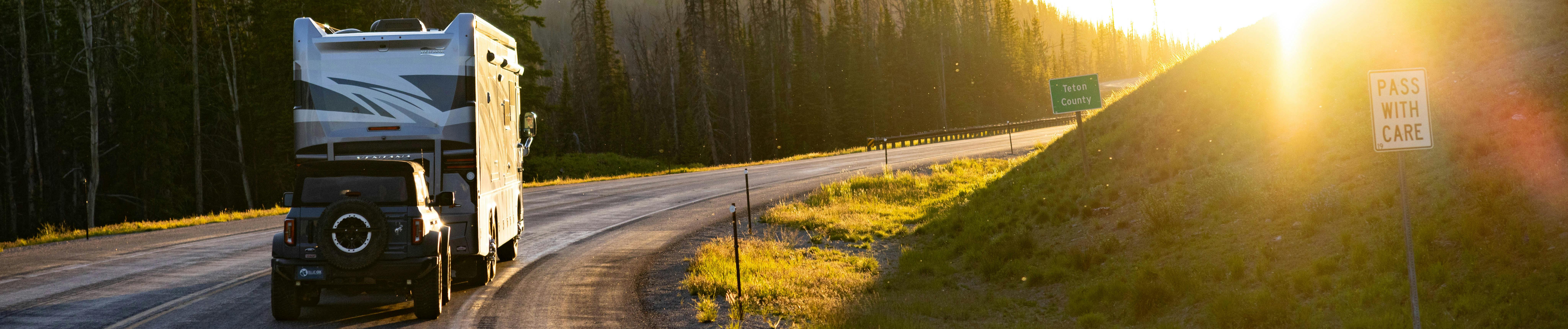 A truck towing a large RV along a winding highway at golden hour, surrounded by pine trees with warm sunlight breaking through — Trademasters flat tow services.